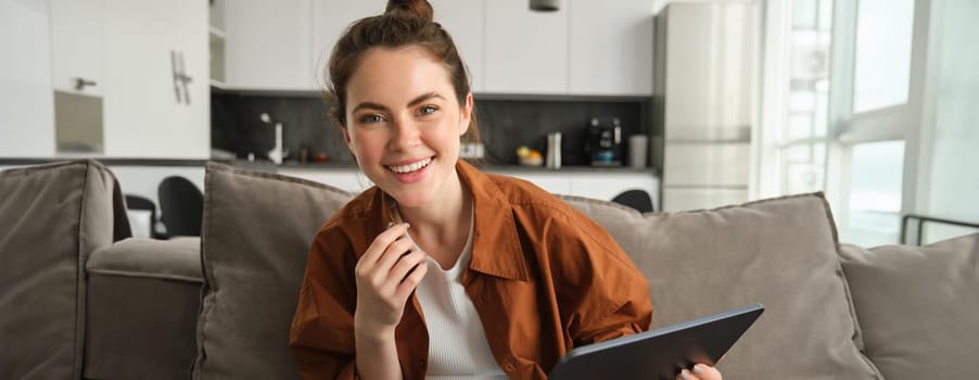 Carefree woman sitting on couch and using digital tablet, laughing and smiling while looking at camera.