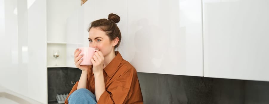 Portrait of young woman with cup of coffee, sits in kitchen and drinks aromatic drink at home, holds tea mug.