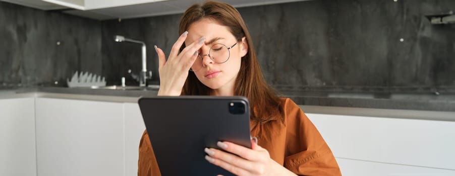 Portrait of young woman feeling unwell, touching head, has headache or migraine, reading on digital tablet in glasses.