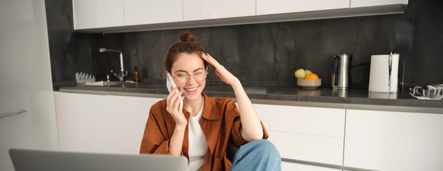 Portrait of beautiful woman in glasses, sitting with laptop in the kitchen, talking on mobile phone, answer a call, laughing and smiling during conversation.