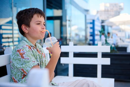 Adorable Hispanic teenage boy drinking healthy smoothie, sitting on a cafe outdoor. Food and drink consumerism.