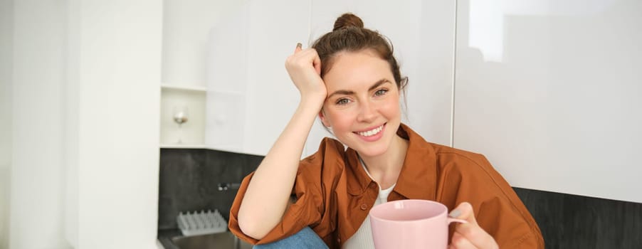 Portrait of woman with cup of coffee in hand, sits in kitchen and smiles at camera. Lifestyle and people concept