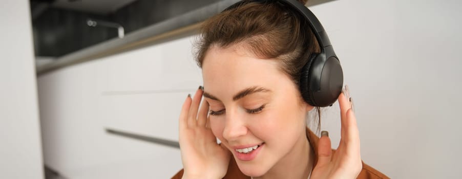 Close up portrait of beautiful brunette woman, listens to music in wireless headphones, has new earphones on her head, smiling and looking happy.