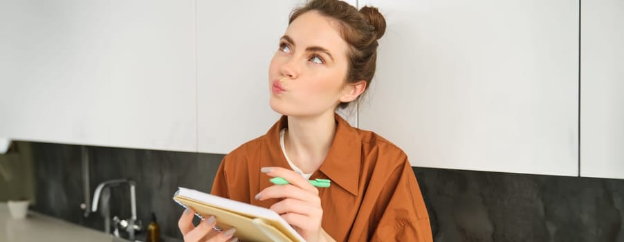 Portrait of cute young woman with notebook, sitting in the kitchen and writing her to do list, making tasks for week, puts notes in schedule or planner.
