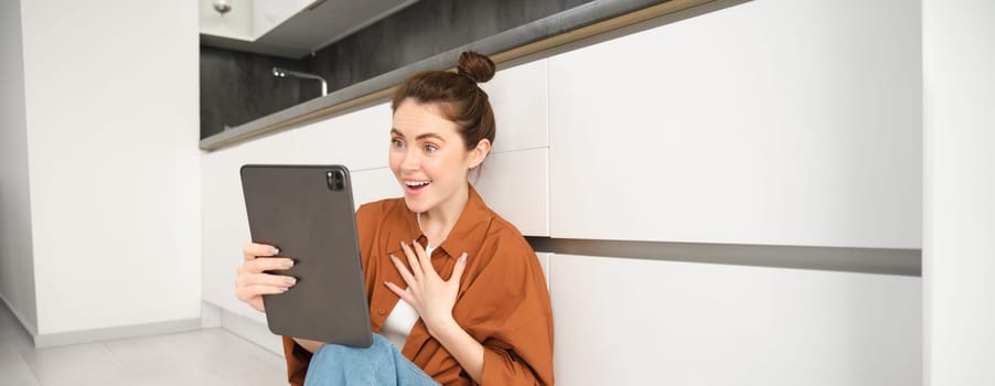 Portrait of friendly smiling woman video chats, connects to online conversation on digital tablet, says hello at camera.