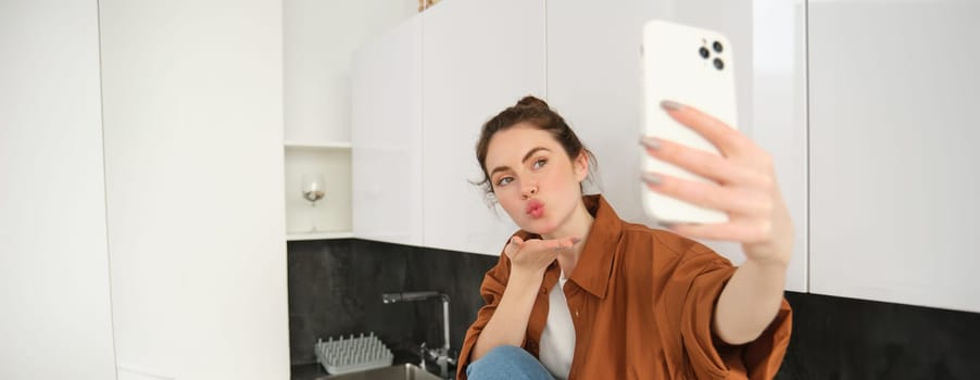 Beautiful young woman, social media influencer takes pictures, records video with smartphone camera, posing on kitchen counter at home, lifestyle blog and people concept.