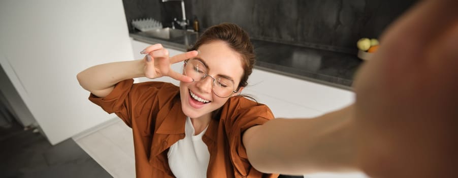 Portrait of carefree young woman in glasses, taking selfie from home, lifestyle blogger making photos in kitchen, laughing and smiling, using smartphone camera.