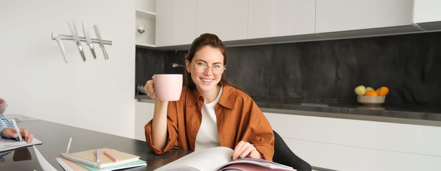 Portrait of young woman, tutor preparing lesson for students, sitting at home, flipping pages of book and drinking tea, student doing homework in kitchen.