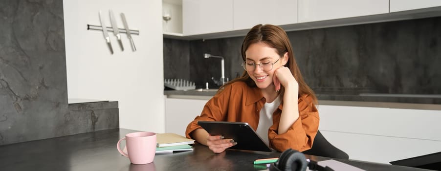 Portrait of young woman working from home, connecting to lesson on digital tablet, reading in glasses, studying remotely in her kitchen, distance learning and education concept.