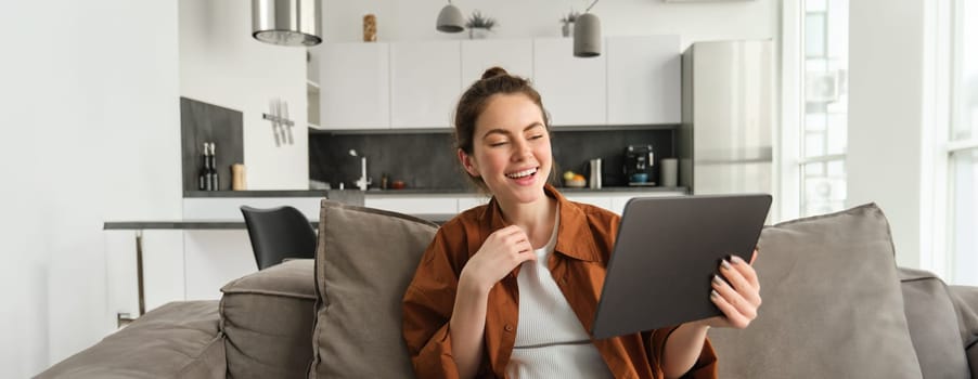 Portrait of woman laughing over funny video on digital tablet, watching movie on her gadget streaming service, sitting on sofa at home, reading e-book.