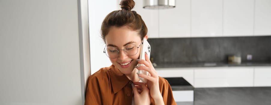 Portrait of lovely young woman answers phone call and smiling, leaning on wall, standing at home and talking on telephone.