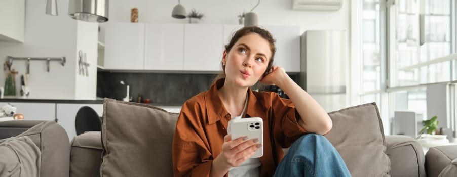 Portrait of cute curious woman, sitting on sofa with smartphone, looks up and thinking, placing an order, online shopping on mobile phone app.