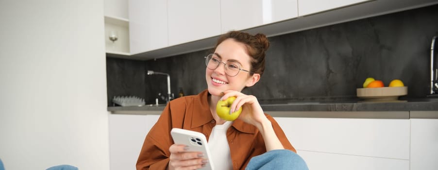 Image of young modern woman, relaxing at home, sitting on chair and using smartphone, eating green apple and smiling.