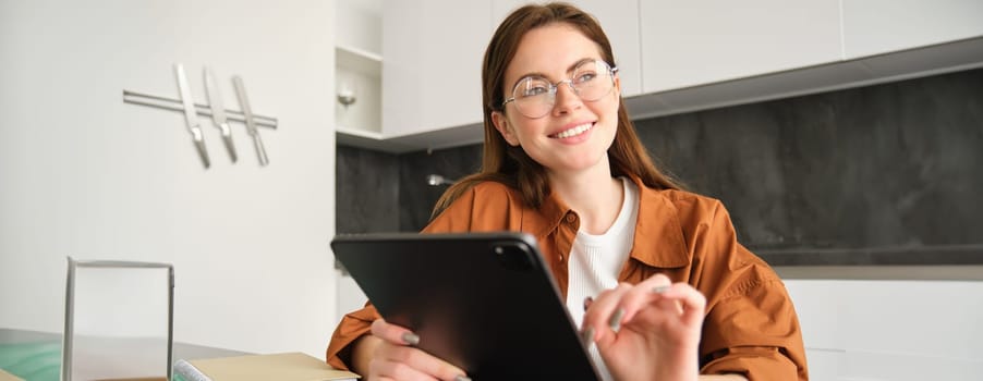 Portrait of young woman working from home, connecting to lesson on digital tablet, reading in glasses, studying remotely in her kitchen, distance learning and education concept.