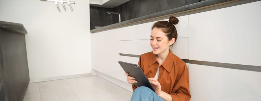 Lifestyle shot of young woman, student using her digital tablet, laughing and smiling, sitting on kitchen floor at home.