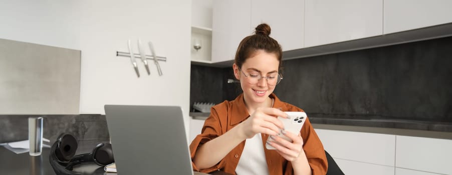 Portrait of young female freelancer, woman working from home with laptop, girl programmer sitting with computer in kitchen, answer message on smartphone.