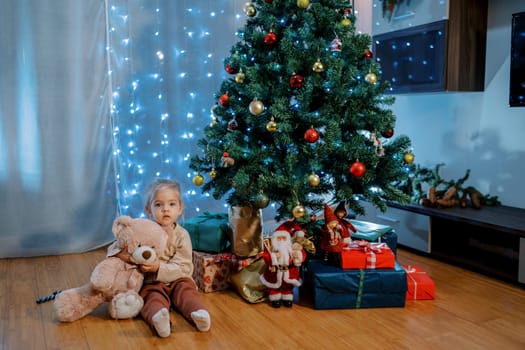 Little girl sits hugging a teddy bear on the floor next to gifts under the Christmas tree. High quality photo