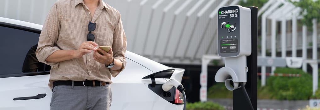 Young man use smartphone to pay for electricity at public EV car charging station at city commercial mall parking lot. Modern environmental and sustainable urban lifestyle with EV vehicle. Expedient