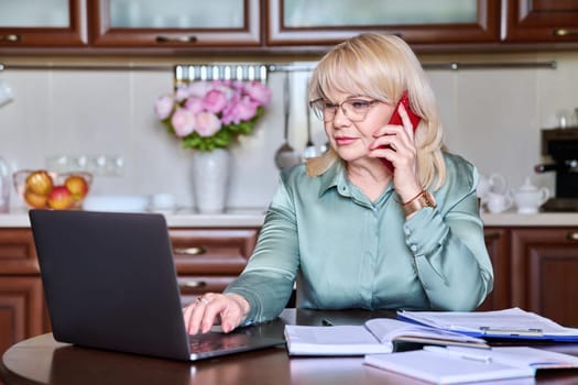 Mature business woman working from home, using laptop sitting in kitchen. Senior 50s female entrepreneur accountant, wearing glasses talking on the phone. Technology virtual online work