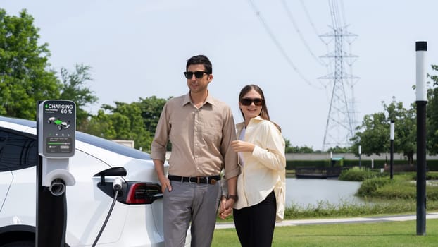 Young couple recharge EV car battery at charging station connected to power grid tower electrical industrial facility as electrical industry for eco friendly vehicle utilization. Expedient
