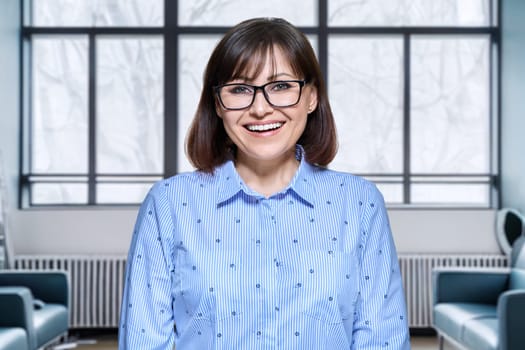 Portrait of mature business woman looking at camera in modern office interior. Positive smiling middle aged female leader manager boss. Job, employee of bank, confidence, career, people concept