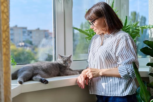 Mature smiling woman looking out window at home with relaxed cat lying on windowsill enjoying warmth of sun. Lifestyle home comfort joy happiness, animals pets, middle aged people concept