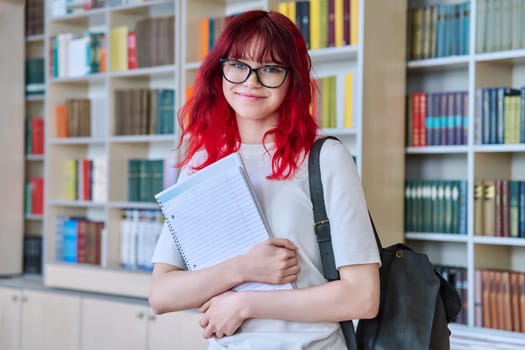 Portrait of teenage female student looking at camera in library. Smiling beautiful girl teenager wearing glasses, holding notebook backpack. College, university, education, knowledge, youth concept