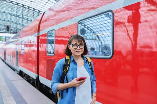 Woman waiting for railway public electric transport on platform of city station. Female with smartphone using banking app to buy online ticket payment, online timetable and route service, technology