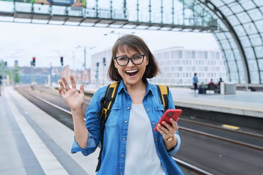 Mature happy woman with smartphone backpack looking at camera inside modern train station building, on platform. Emotion joy happiness, transportation, journey, trip, middle age people concept