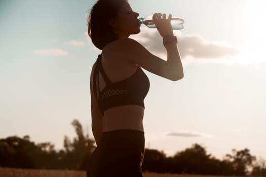 A young girl takes a break during an intense workout and jogging and drinks refreshing cool water from a bottle, a woman trains outdoor in the evening at sunset.