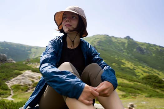 A happy young girl in a hat and windbreaker sits on a rock and enjoys an amazing view with mountains, hills and forest during her journey, hiking in the wild. A woman spends an active lifestyle.