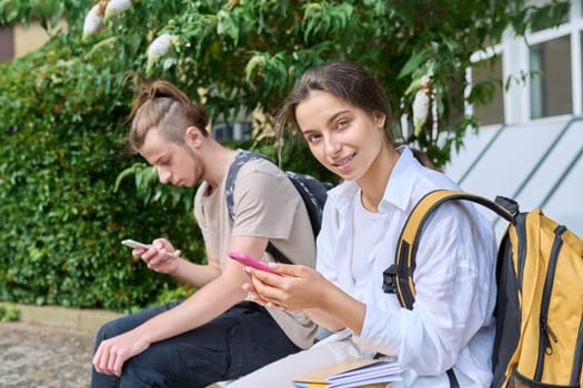 Teenage high school students, guy and girl, sitting on bench with backpacks, near academic building, using smartphones. Adolescence, youth, education, lifestyle, technology concept