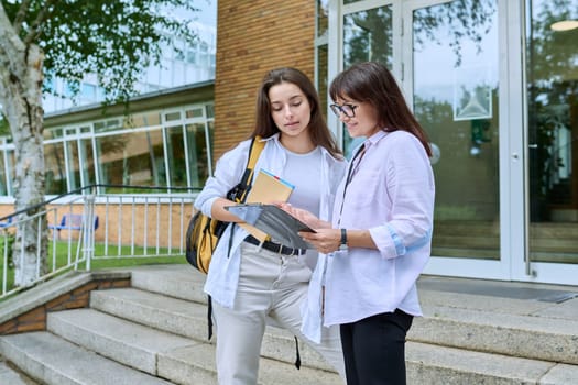 Teenage girl high school student with backpack talking to female teacher, mentor, coach, standing outdoors on steps of educational building. Adolescence, education, knowledge, communication