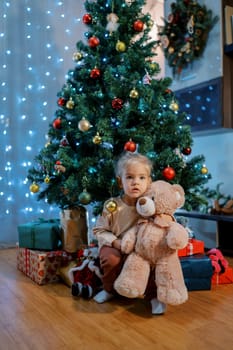 Little girl sits on gifts under the Christmas tree, hugging a teddy bear. High quality photo