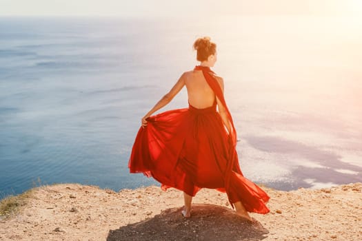 Side view a Young beautiful sensual woman in a red long dress posing on a rock high above the sea during sunrise. Girl on the nature on blue sky background. Fashion photo.