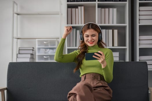 Woman wearing headphones plays video game on smartphone while relaxing on sofa in home lifestyle..