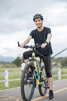 A happy and handsome Asian man in sportswear and a bike helmet, enjoys listening to music through his earbuds while riding a bike on the country roads on the weekend..