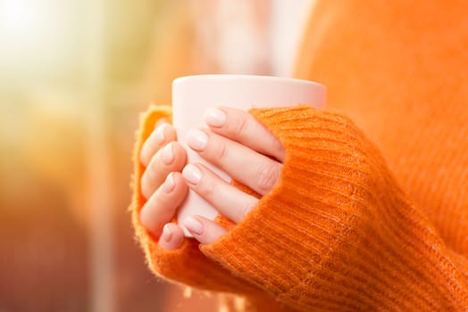 A woman drinks coffee at home with the sunrise through the window. Close up of womans hands are holding hot coffee cup near window at home