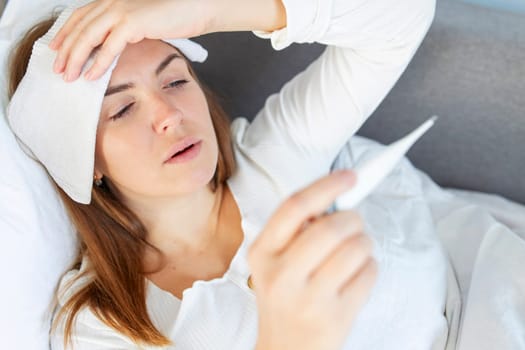 Woman with flu or covid-19 lying in bed, she is measuring her temperature with a thermometer and touching her forehead. Sick woman.
