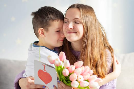 Happy Mother's Day! Happy little boy congratulating his mom and giving her flowers tulips and greeting card with red heart drawn.