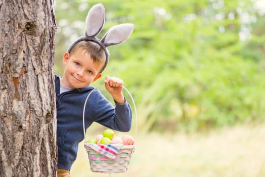 Happy child looks out of a tree holding basket full of colorful easter eggs after egg hunt at spring time. Happy Easter day.