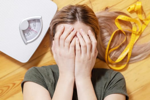 Upset and sad woman covering face with hands while lying on wooden floor with white scales and measuring tape.