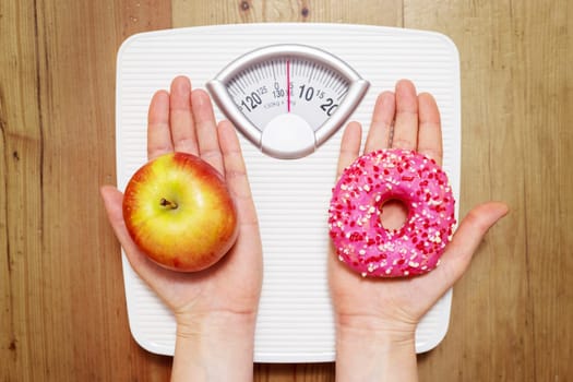 Diet concept. Female hands hold sweet donut and red apple against the background of scales, choice between healthy unhealthy food dessert. Top view.