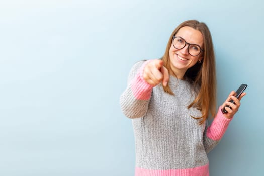 Portrait of happy brunette woman in glasses holding smartphone and pointing her index finger at the camera on blue background.