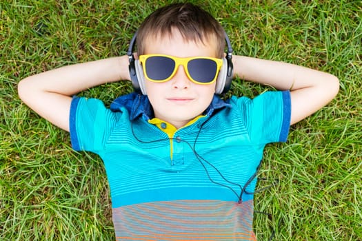Happy boy in sunglasses lying on the grass and listening to music in headphones on a sunny day.