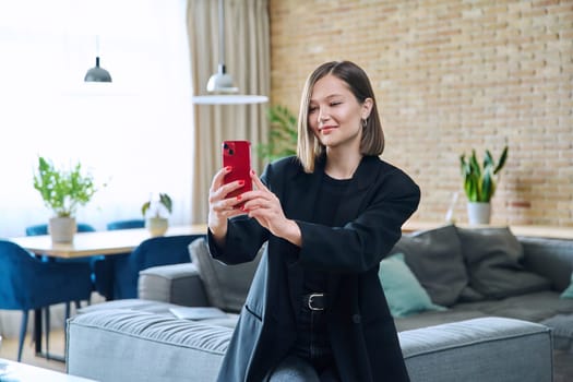 Young smiling joyful happy woman sitting on the sofa in the living room having a video call chat conference using a smartphone. Communication, leisure, home, lifestyle, youth, technology concept