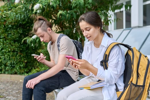 Teenage high school students, guy and girl, sitting on bench with backpacks, near academic building, using smartphones. Adolescence, youth, education, lifestyle, technology concept