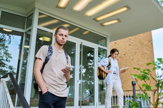 Hipster guy smiling teenager student 18, 19 years old with backpack smartphone looking at camera, standing outdoor educational building. Youth, education, lifestyle, technology