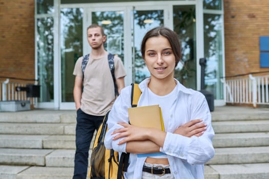 Portrait of high school student, smiling girl with backpack looking at camera, outdoor on steps of educational building. Adolescence, 15, 16, 17 years old, lifestyle, education concept