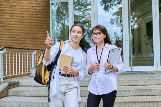 Teenage girl high school student with backpack talking to female teacher, mentor, coach, standing outdoors on steps of educational building. Adolescence, education, knowledge, communication